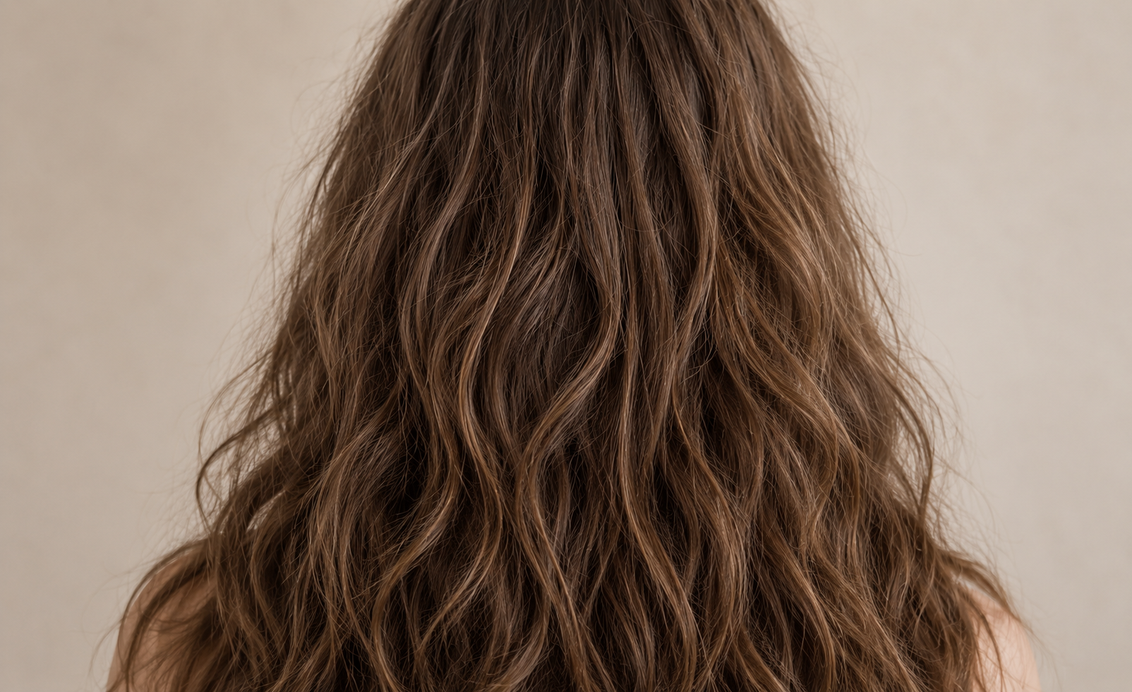 Close-up view of frizzy hair with natural texture, flyaways and soft waves against a neutral beige studio background.