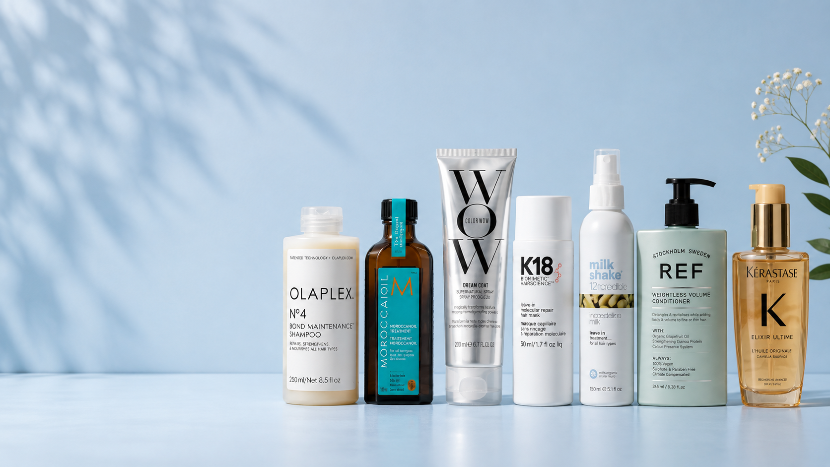 Six haircare products from different brands are lined up on a light blue surface against a soft blue background with leaf shadows. The products include Olaplex, Morphe, WOW, K18, Ref, and Kérastase.